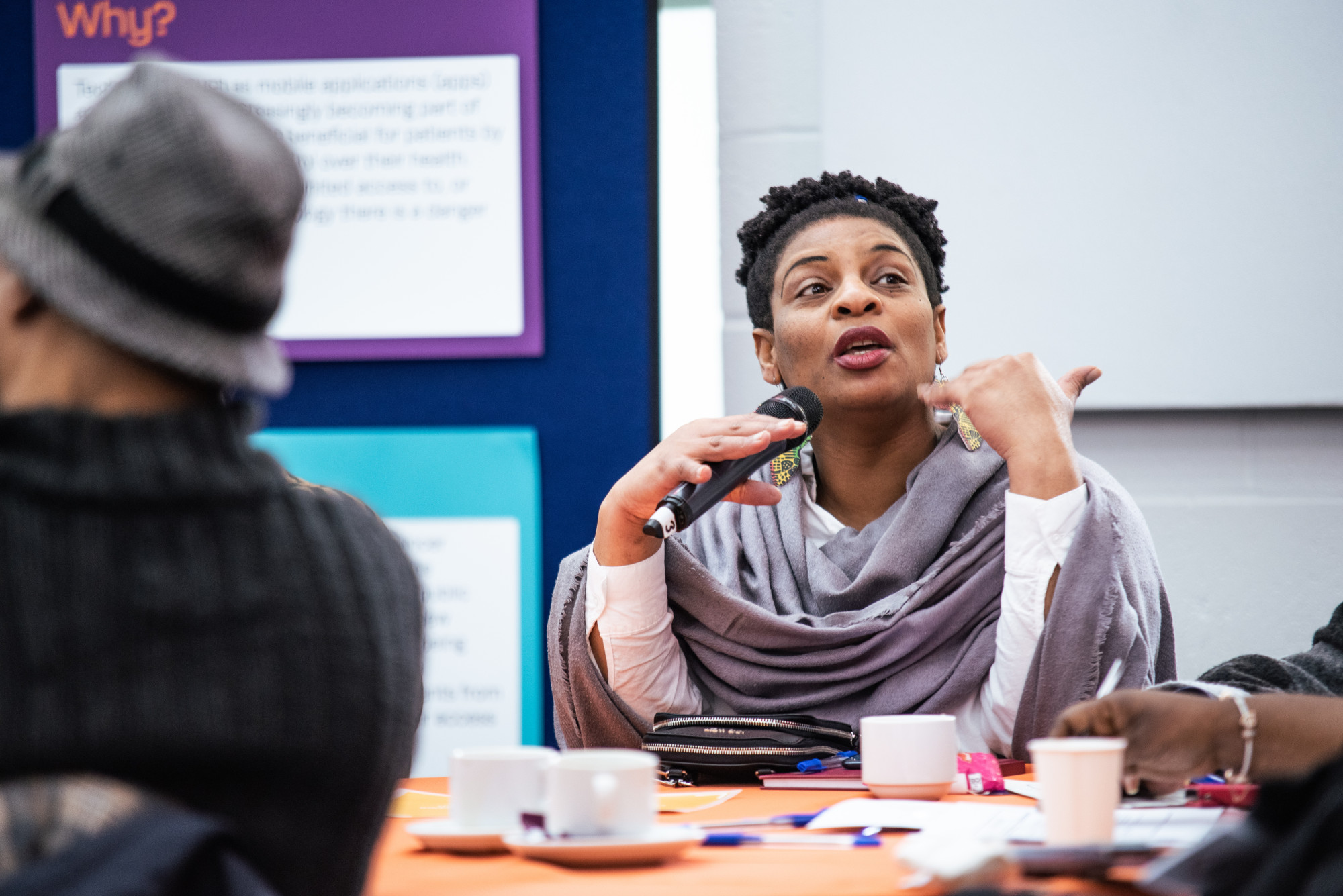 A person speaks into a microphone while sitting at a table with cups, surrounded by others.