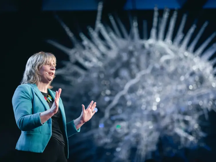 A woman in a teal blazer speaks on stage with her hands raised; a large, abstract, spiky sculpture is in the background.