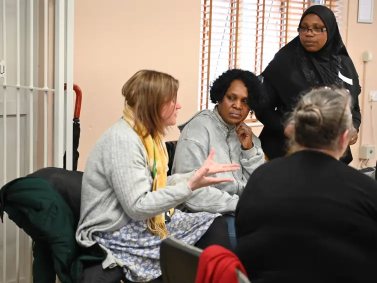 Four women engaged in a serious conversation in a small room. One woman gestures expressively while others listen.