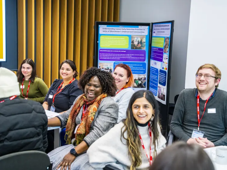A group of people seated in a room, engaged in a discussion or meeting. Posters or presentation boards with text and images are visible in the background.