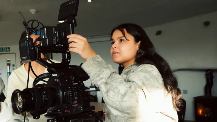 A young woman with long dark hair operates a professional video camera indoors, adjusting its monitor while looking attentively at the equipment. Sunlight shines on her face and sleeve.