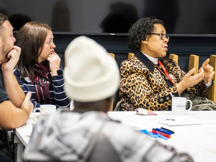 A group of people sitting at a table. One of the people is wearing a leopard print jacket and is talking to someone off camera, gesticulating with their hands.
