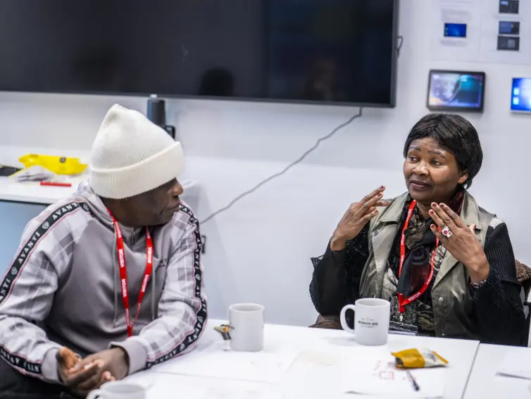 The environment is an office. Two people talking to each other, the woman is gesticulating as she talks. She's a Black African woman wearing a brown jacket over a black shirt and her hair is in bob. The other person, a Black man wearing a white beanie and grey tracksuit, is listening to the woman.