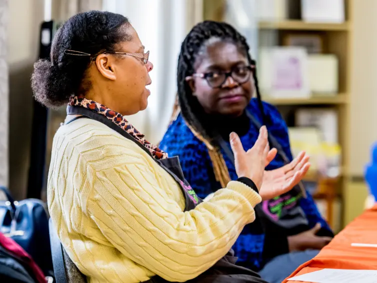 Two people sitting at a table in discussion. One is gesturing with their hand while speaking. Both are wearing aprons, and there are papers and a bag on the table. The room has bookshelves in the background.