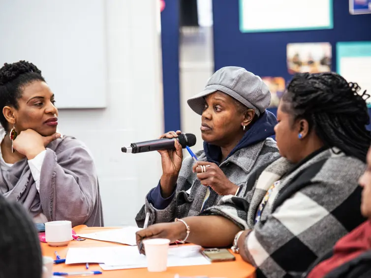A group of people around an orange table engaged in discussion. A woman in a gray cap speaks into a microphone while others listen. Papers, cups, and pens are on the table, with a bulletin board in the background.