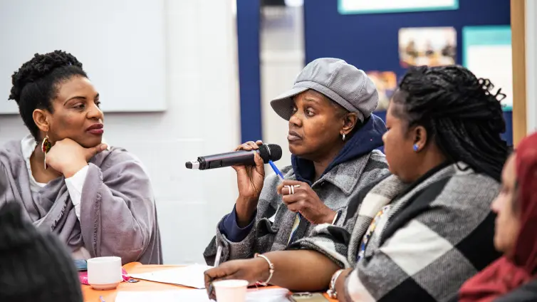A group of people around an orange table engaged in discussion. A woman in a gray cap speaks into a microphone while others listen. Papers, cups, and pens are on the table, with a bulletin board in the background.