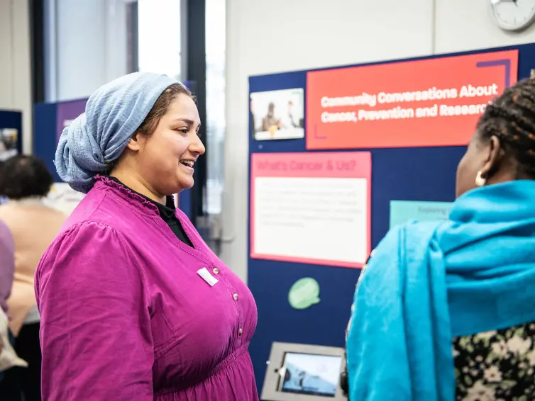 A woman in a purple dress and headscarf smiles while talking to another woman in a blue headscarf and patterned jacket. They are at an event with displays about cancer awareness and research.