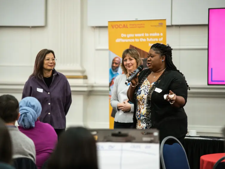 Three women stand and speak at a presentation. One holds a microphone. Attendees sit and listen. A yellow poster and a screen are visible in the background.
