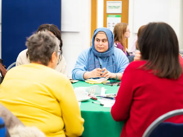 A group of people sitting around a table covered with a green tablecloth, engaged in discussion. The table has papers, pens, and cups on it. A woman wearing a blue hijab and sweater is attentively listening, with her hands clasped together. Other participants, wearing colorful clothing, are seated with their backs partially visible. The setting appears to be a community or workshop space, with posters and signs on the walls in the background.