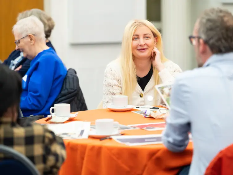 A group of people sitting around a round table with a bright orange tablecloth. One person is smiling and looking to someone in front of her.