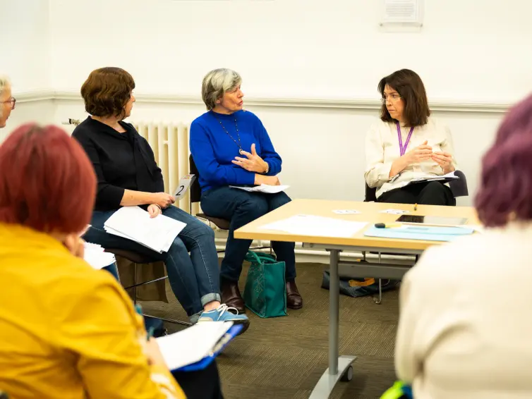A group of people sitting in a room. One person with an electric blue jumper is saying something to a woman wearing a white shirt and purple lanyard.