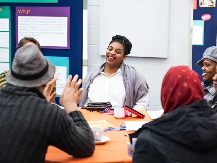 People sitting around a table, engaged in a friendly discussion. A woman in the center is smiling, and others are actively participating.
