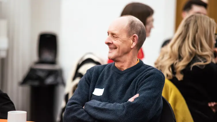 A person smiling and looking away from the camera, their arms are crossed and they're sitting next to a table with orange cover.