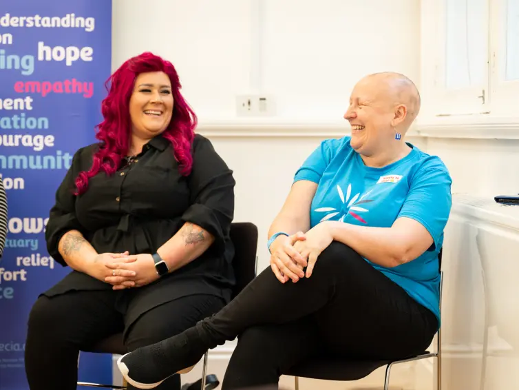 Two women sitting on chairs against a bright blue wall, appearing relaxed and engaged with each other. They're smiling and laughing.